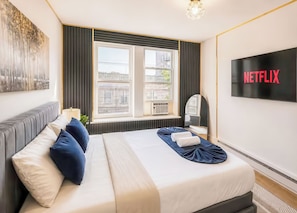 A hotel room with a bed, a TV screen displaying 'NETFLIX,' a window with blinds, and a view of buildings outside. - 20 Mins to Times Square Skyline Views (West New York)