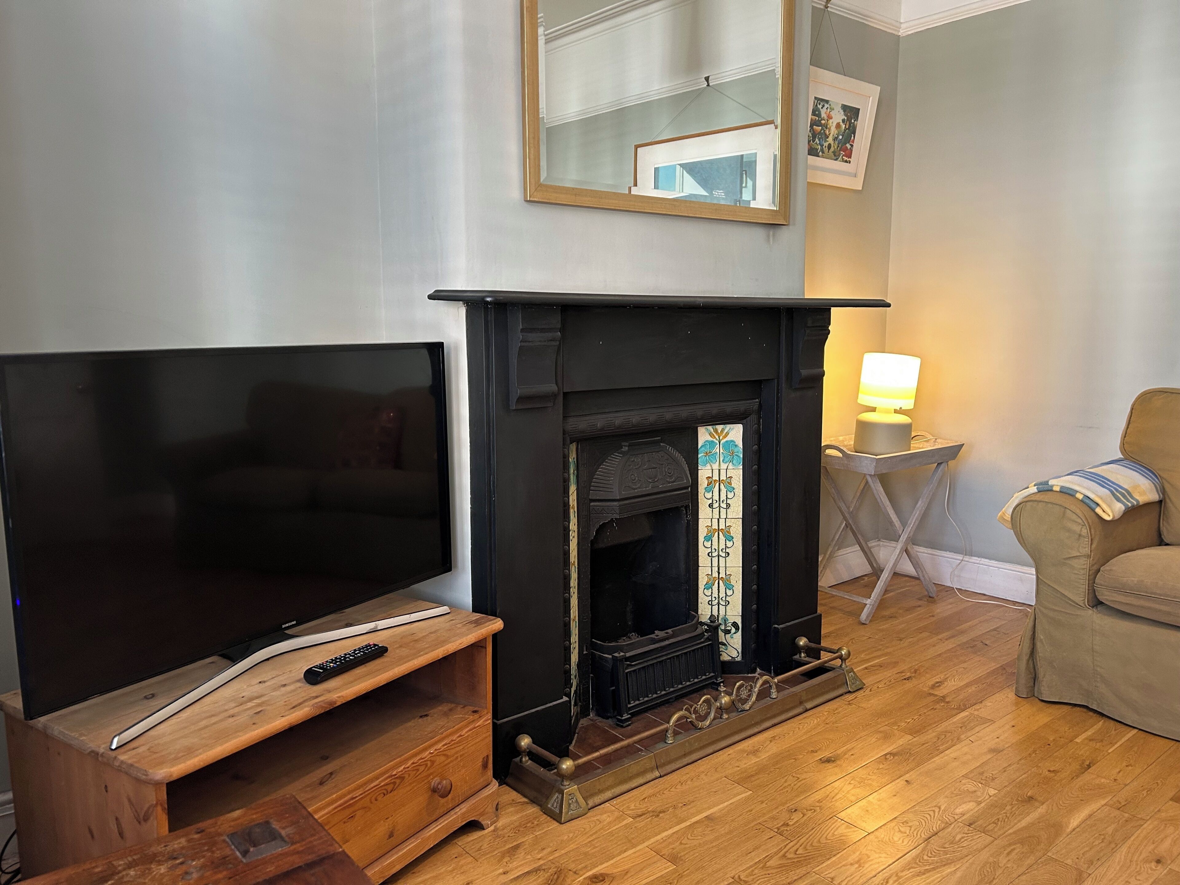 Ein Wohnzimmer mit Fernseher auf einem Holzständer, Kamin mit dekorativen Kacheln, einem kleinen Tisch mit Lampe und einem Sofa.