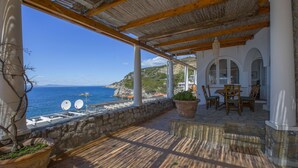 Una terraza costera con vistas al mar, muros de piedra y un área de comedor con sillas y una mesa.