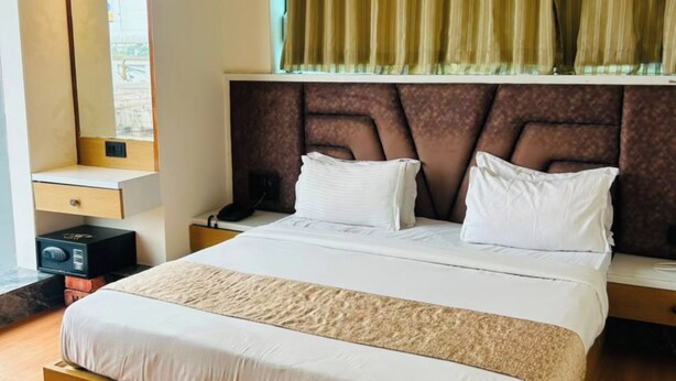 A modern hotel room with a large bed, a wooden headboard, a bedside table, a nightstand, a mirror, and a window with curtains. - KOKO Regency Hotel & Hospitality (Gandhinagar)