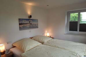 Ein Schlafzimmer mit zwei Betten, einem Gemälde von einem Leuchtturm, einem Fenster und einem Nachttisch mit einer Lampe.