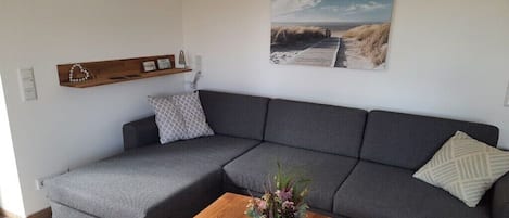 Un salon moderne avec un canapé d’angle gris, une table basse en bois agrémentée d’un centre de table floral, une étagère murale décorée de divers objets et un tableau sur le thème de la plage.