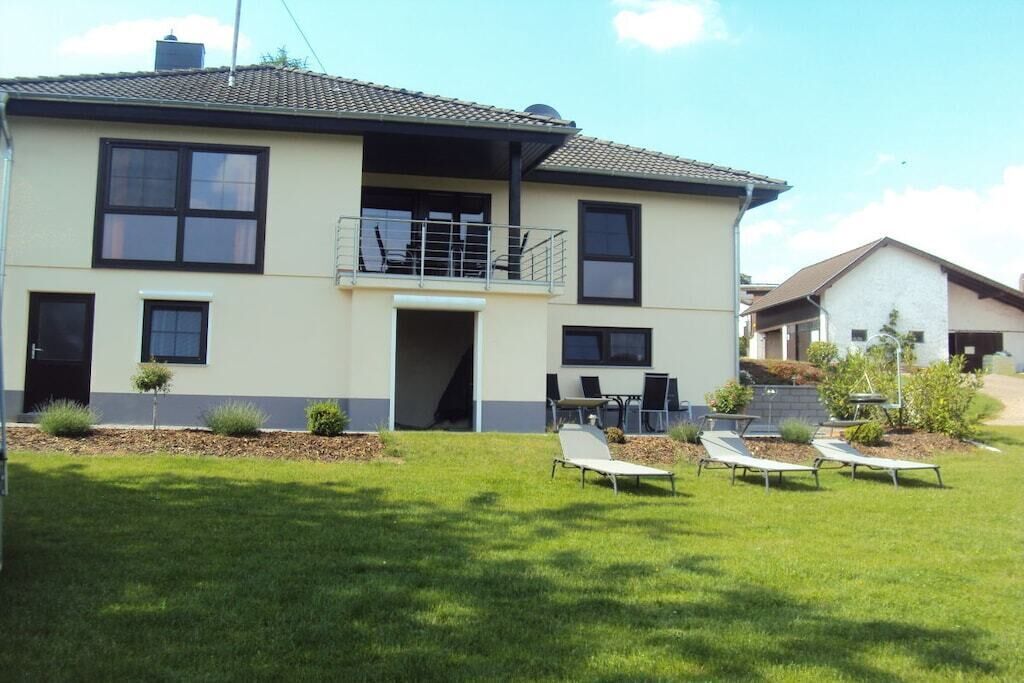 A two-story house with a balcony, a lawn with sun loungers, and a small garden area.