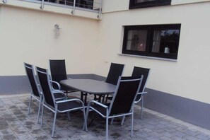 An outdoor patio with a round table and four chairs, a window with a black frame, and a building with a light-colored exterior. - Holiday home with 1 bedroom (Mettlach-Tünsdorf)