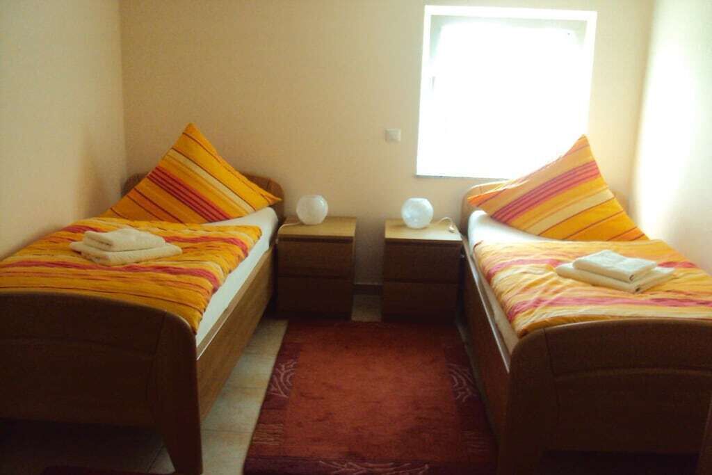 A room with two single beds, each with a striped bedspread and pillows, a nightstand with a lamp, and a window providing natural light.