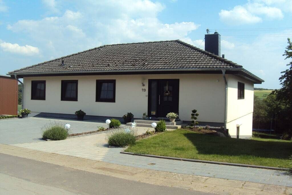 Einbungalow mit Ziegeldach, Hausnummer 19, mit Eingangstür, Fenstern und einem kleinen Vorgarten mit Pflanzen und einem Weg.