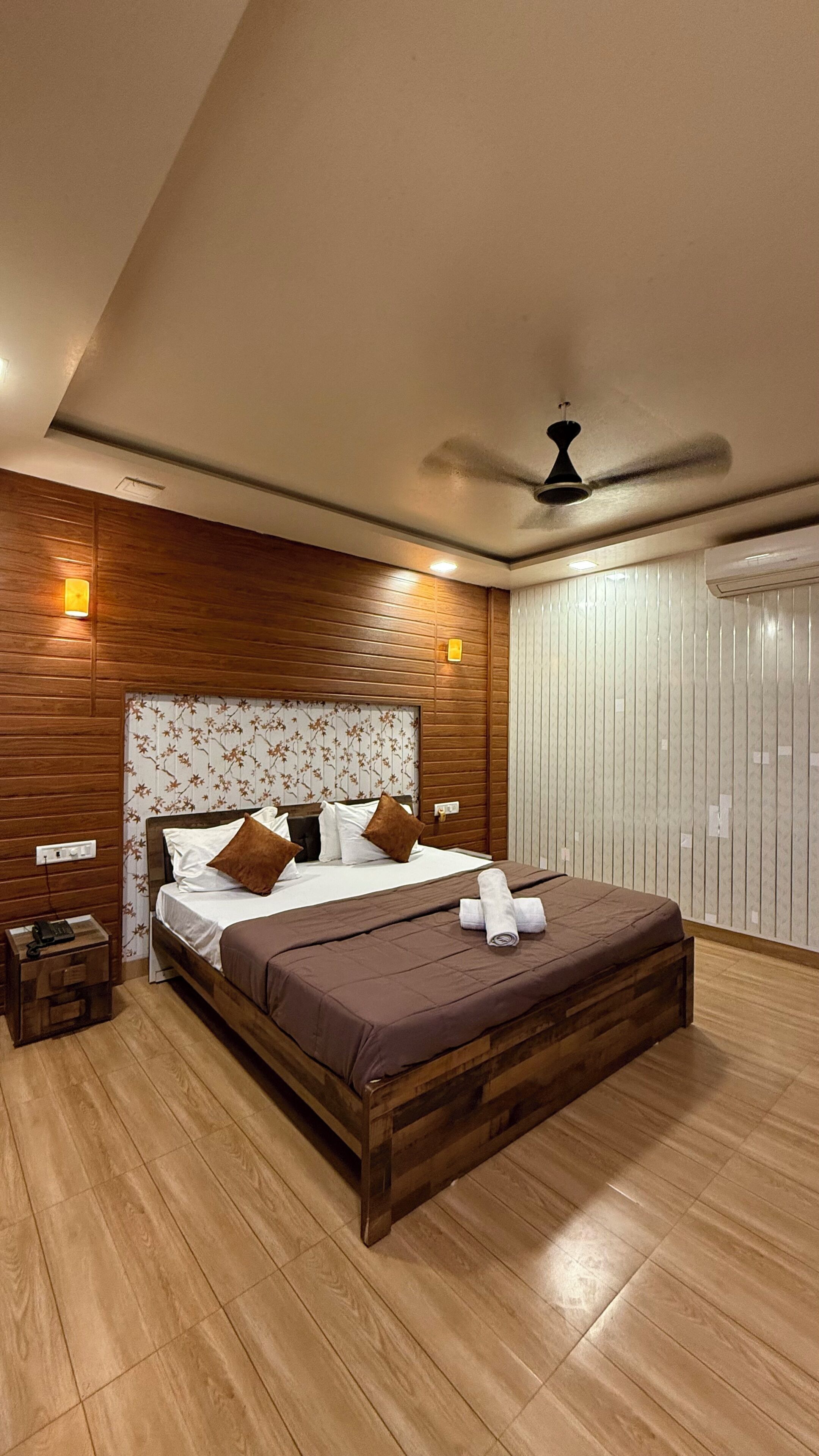 Une chambre moderne avec une tête de lit en bois, un lit avec une literie marron, une table de chevet avec un téléphone et un ventilateur de plafond.