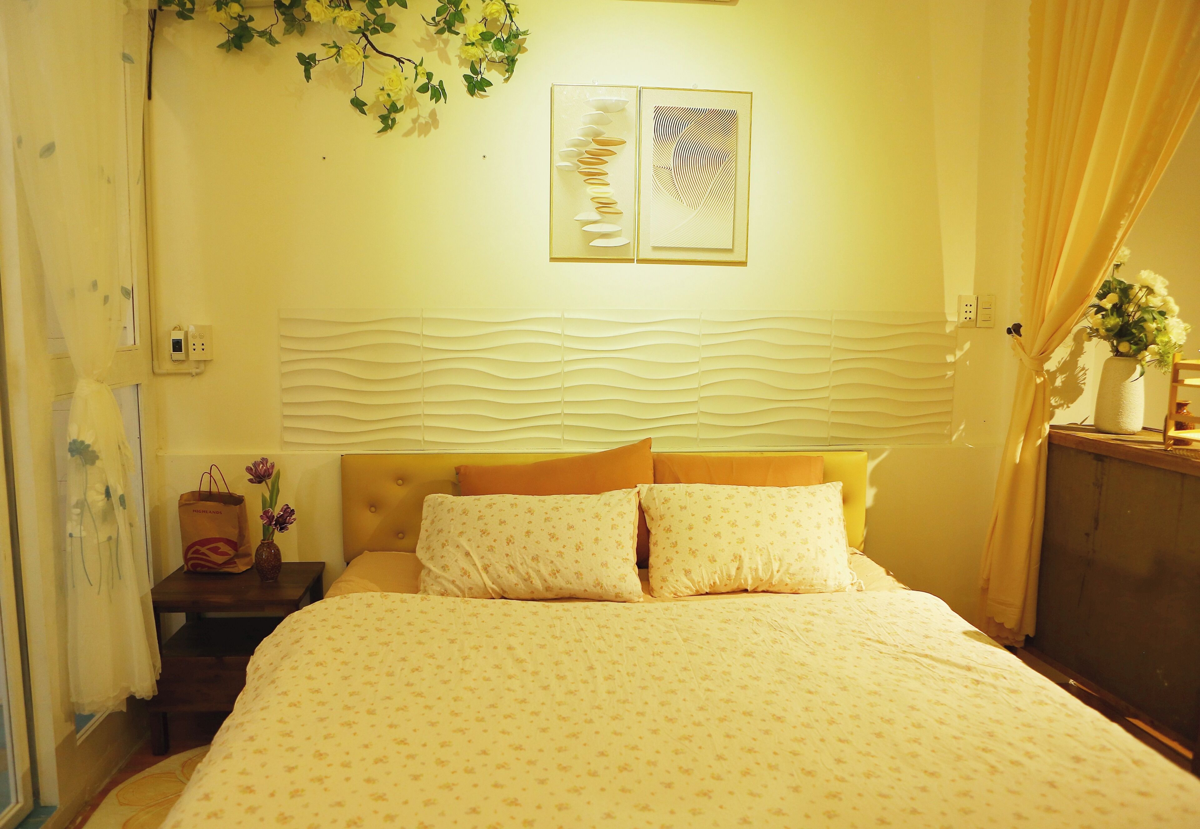 A hotel room with a bed, a nightstand, a vase with flowers, and a wall with decorative elements.