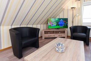 Ein Zimmer mit schrägem Dach, einem Fernseher, zwei schwarzen Stühlen, einem Holztisch und einem kleinen dekorativen Korb.
