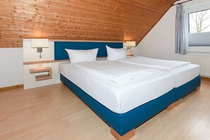 A bedroom with a wooden sloped ceiling, a bed with white bedding, a blue headboard, and a window with curtains. - holiday apartments "Deichblick" (Esens-Bensersiel)