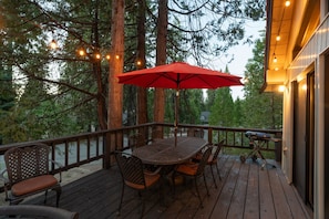 An outdoor deck with a table and chairs, surrounded by tall trees and string lights. - Your cozy home away from home in Shaver Lake! (Shaver Lake)