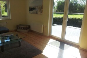 Een kamer met een glazen tafel, een bank, een radiator, een mandje met de tekst 'HOME' en een uitzicht door een glazen deur.