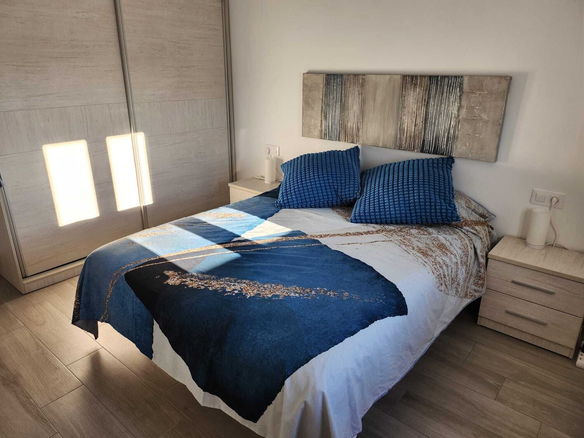 Quarto moderno com cama grande, mesa de cabeceira, luminária na parede e janela com persianas.