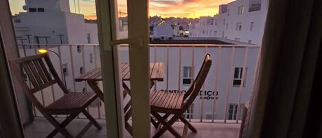 Un balcon avec deux chaises en bois et une table, d’où l’on peut admirer la vue sur la ville au coucher du soleil.