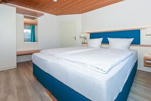 A bedroom with a bed, a window with curtains, a door, and a wooden ceiling. - Holiday apartments "Flut" (Esens-Bensersiel)