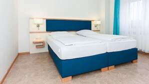En dubbelsäng med en blå sänggavel och vita sängkläder i ett hotellrum med ett kaklat golv och ett fönster med blå gardiner.