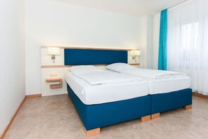 Cama doble con cabecera azul y ropa de cama blanca en una habitación de hotel con piso de baldosas y una ventana con cortinas azules.