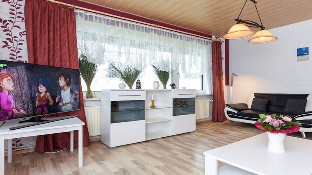 Ett modernt vardagsrum med en tv, vita möbler, en svart soffa och ett fönster med gardiner.
