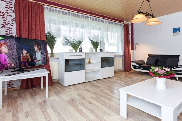 Una sala de estar moderna con televisor, muebles blancos, un sofá negro y una ventana con cortinas.