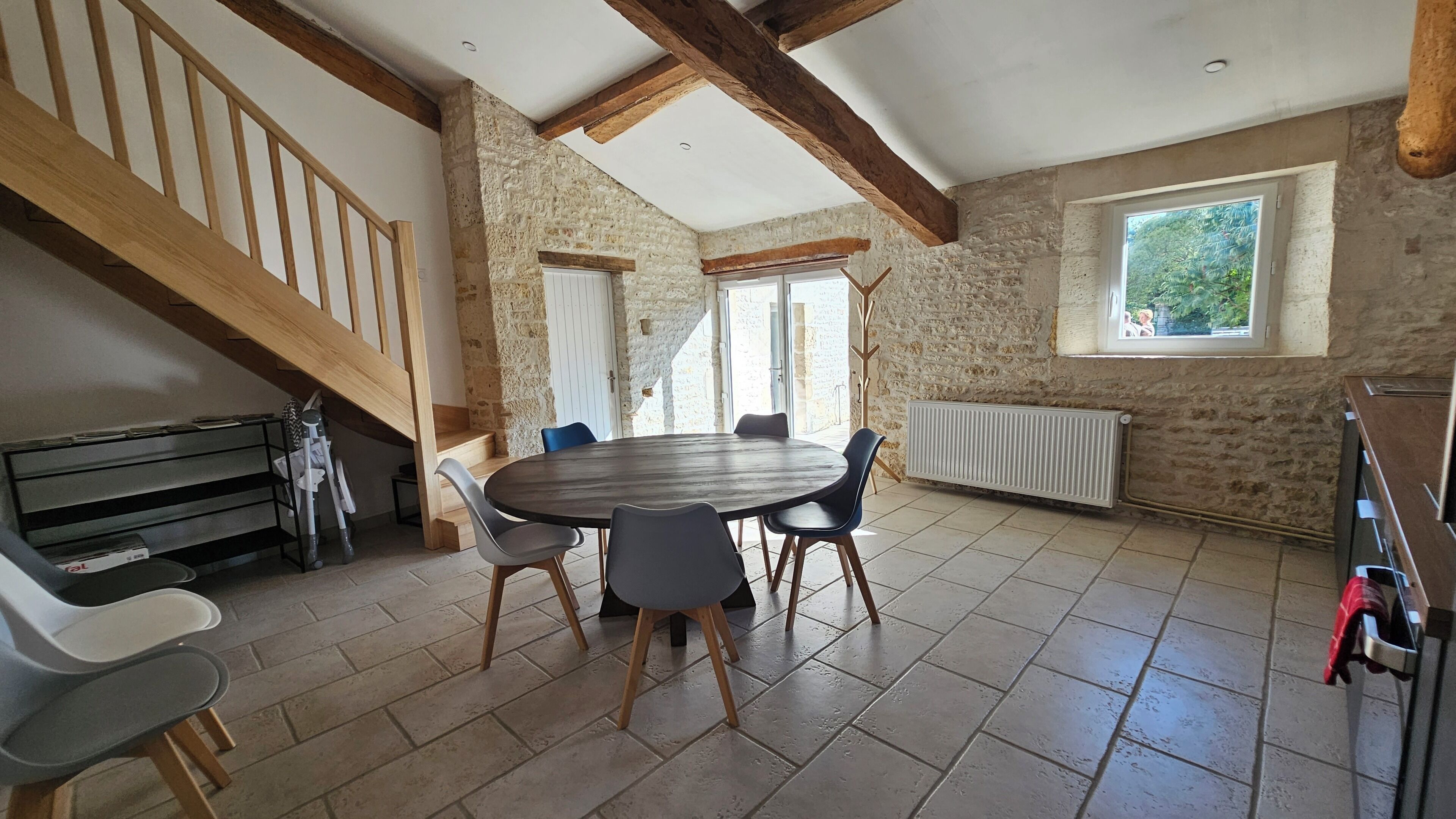 A room with a stone wall, a wooden staircase, a round table with chairs, and a window with a view of greenery.