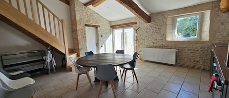 A room with a stone wall, a wooden staircase, a round table with chairs, and a window with a view of greenery.