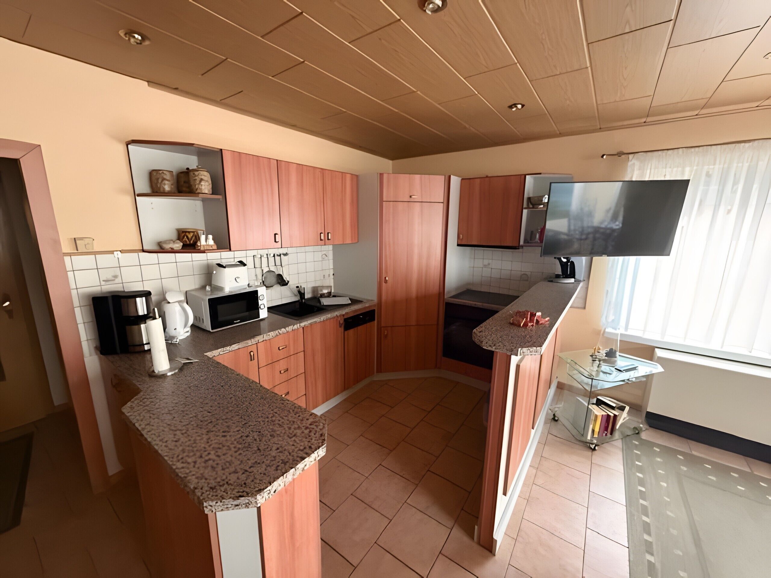 Een moderne keuken met houten kastjes, een magnetron, een koffiezetapparaat en een tv boven de aanrecht.
