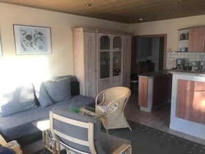 Un espace de vie comprenant un canapé, un fauteuil, un meuble avec des portes en verre, et un coin cuisine avec un plan de travail et des appareils électroménagers.