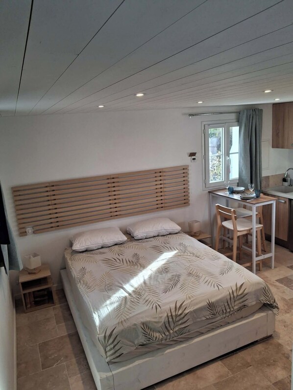 A bedroom with a bed, a wooden headboard, a small bedside table, a window with curtains, and a kitchenette area with a sink and chair.