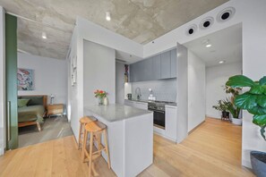 A modern kitchen with grey cabinets, a central island with stools, and a dining area with a sofa and a painting. - Sunlit 2BD Apartment with Parking, Brunswick (Brunswick)
