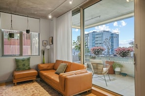 A modern living room with a leather sofa, a small table, and a chair. There is a large window with a view of buildings and a balcony with potted plants. - Sunlit 2BD Apartment with Parking, Brunswick (Brunswick)
