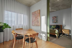 A room with a wooden dining table, chairs, a bed with a green pillow, a bedside table with a lamp, and a painting on the wall. - Sunlit 2BD Apartment with Parking, Brunswick (Brunswick)