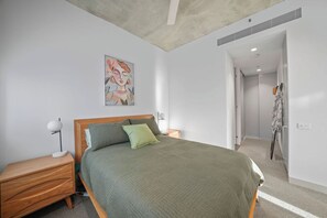Un dormitorio con cama, mesita de noche, lámpara, un cuadro en la pared y una puerta que da a otra habitación.