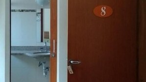 A room with a wooden door marked with the number 8, an air conditioning unit above, and a glimpse of a kitchen area through an open door.