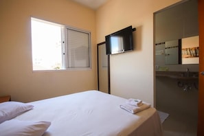A hotel room with a bed, a television mounted on the wall, a window with blinds, and a bathroom with a mirror and sink. - Hotel Paulista (São José do Rio Pardo)
