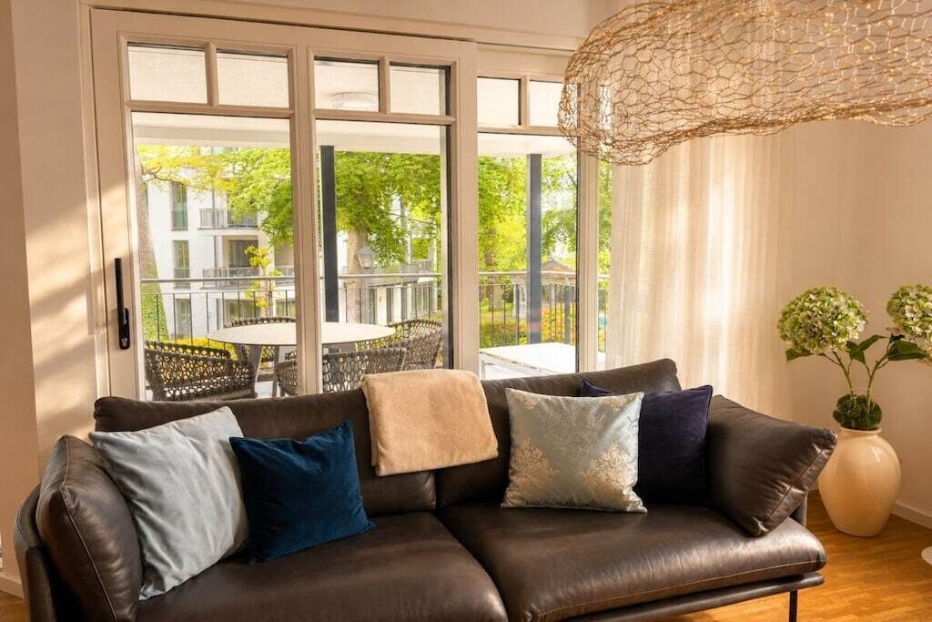 Un canapé en cuir marron agrémenté de plusieurs coussins, une grande fenêtre donnant sur un patio extérieur et un luminaire suspendu.