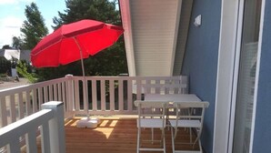 Een houten terras met een witte reling, een rode parasol en een witte tafel met stoelen.