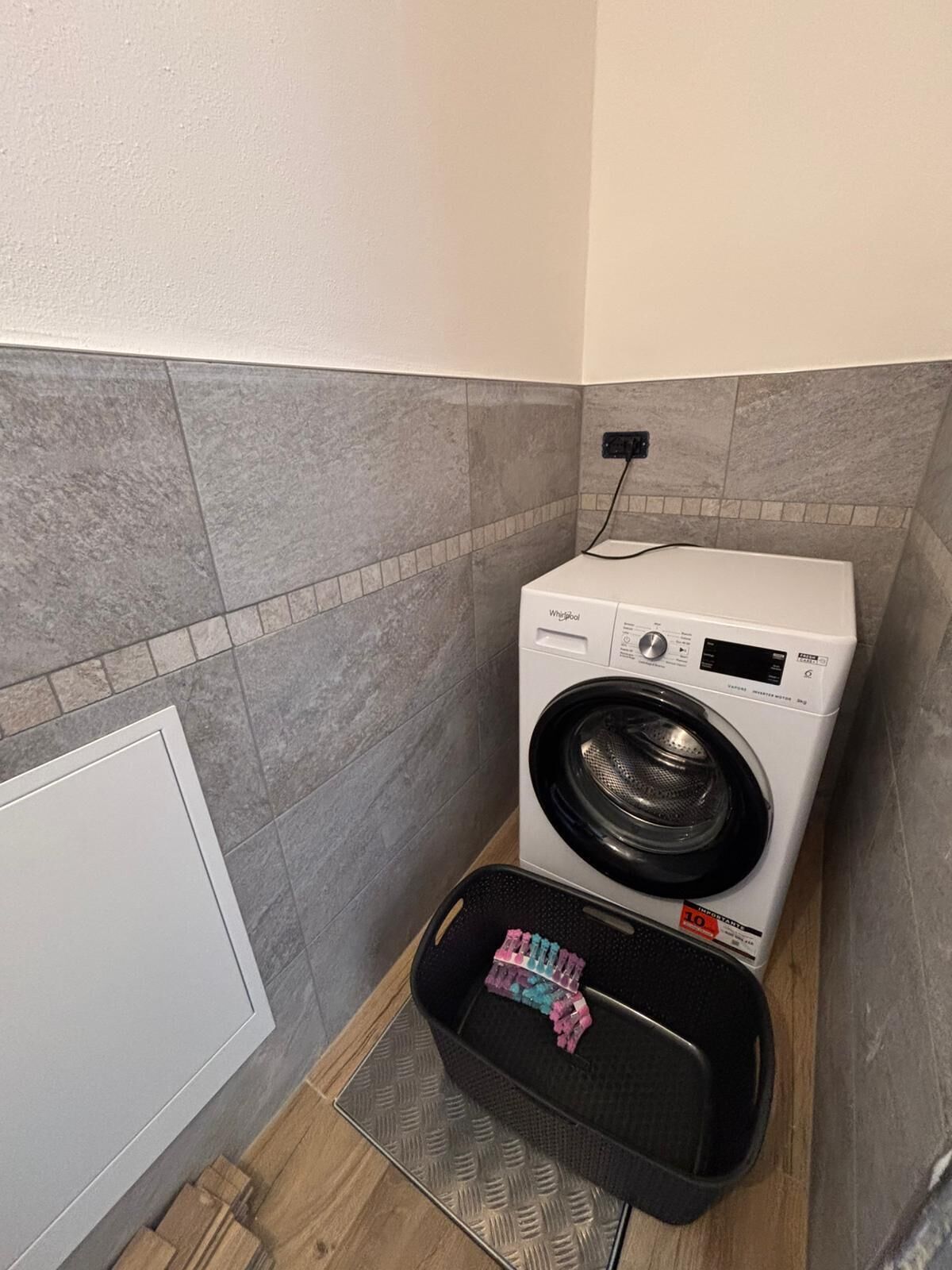 Uma lavanderia compacta com máquina de lavar de abertura frontal, um cesto preto pequeno com peças coloridas e paredes revestidas de azulejos cinzas.
