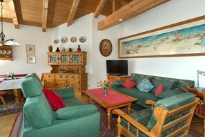 Ein Wohnzimmer mit einem grünen Sofa, Holzstühlen, einem Holzkabinett, einem Fernseher und einem Gemälde an der Wand.