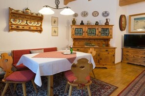Ein Essbereich mit einem Tisch für zwei Personen, Holzkästen, einer roten Tischdecke mit Muster und einer Vitrine mit Dekorationsgegenständen.