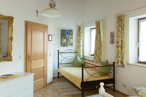 A bedroom with a single bed, a wooden door, a white dresser, a window with curtains, and a wall-mounted light fixture. - Christine Penners 2 - "Kampenwand" (Aschau i. Chiemgau)