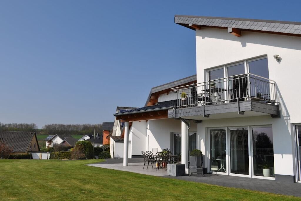 A modern two-story residential building with a balcony, large windows, and a well-maintained lawn.