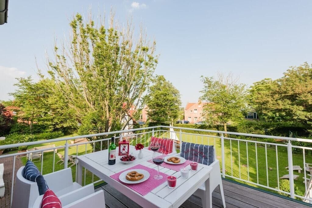 An outdoor dining area with a table set for two, surrounded by a white railing, overlooking a well-maintained garden and residential buildings.