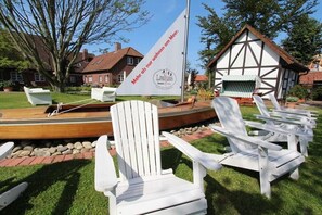 Área de jardim com cadeiras Adirondack brancas, um barco de madeira e uma casa com fachada branca e marrom.