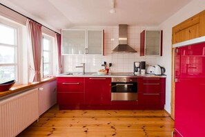 A kitchen with red cabinets, a stainless steel oven, and a window with curtains. - Holiday apartment with 2 bedrooms (Krummhörn - Greetsiel)