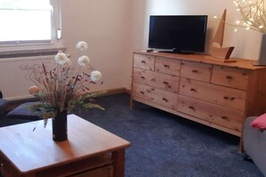 Quarto com cômoda de madeira, televisão em cima e vaso com flores secas sobre uma mesinha.