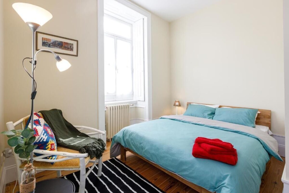 A bedroom with a bed, a chair, a floor lamp, a window, and a radiator.