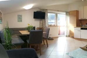 Ein Wohnzimmer mit einer Couch, einem Esstisch, Stühlen, einem Fernseher und einem Fenster mit Vorhängen.