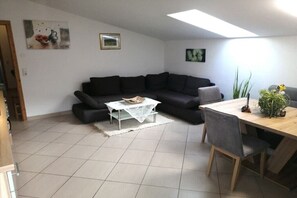 Ein Wohnzimmer mit einem dunkelgrauen Sofa, einem kleinen weißen Couchtisch und einem Holzespeisetisch mit Stühlen. An den Wänden hängen zwei Bilder und auf dem Esstisch steht ein Topf mit einer Pflanze.