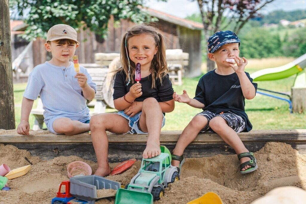 Three children sitting on a wooden bench outdoors with sand toys and a green toy vehicle.