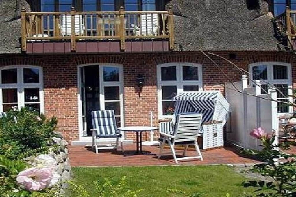 A brick house with a balcony, a patio with chairs and a table, and a garden with flowers.